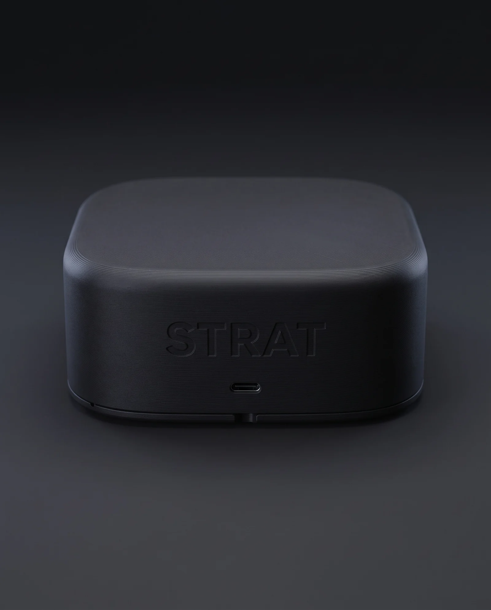 Spatele bazei, de aproape: logo-ul STRAT gravat fin în PLA-ul mat, portul USB-C dedesubt și crestătura din muchia de jos prin care se deschide capacul.